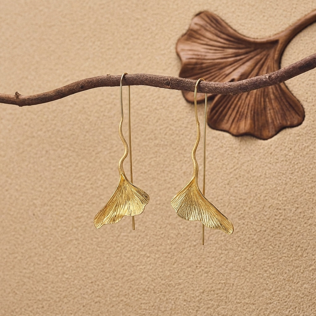 Luxurious gold-plated Ginkgo biloba leaf earrings laid flat on a dark stone surface